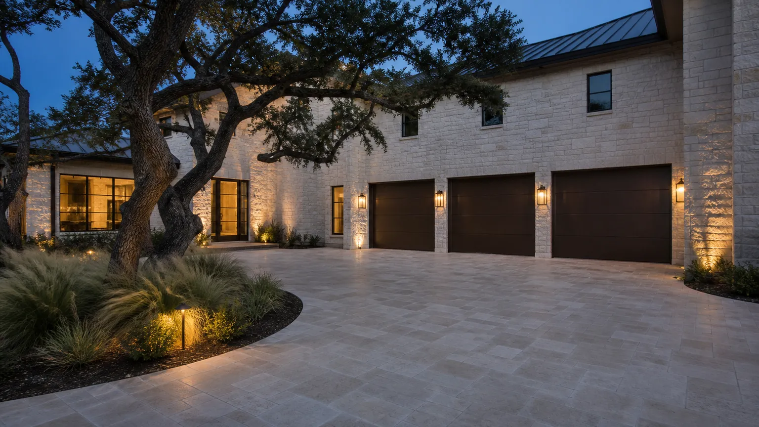 Limestone motor court at dusk with bronze garage doors and low landscape lighting